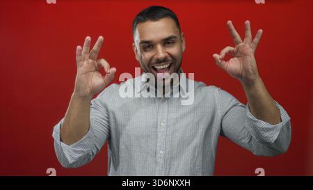 Uomo che fa a mano il segno con entrambe le mani e sorridente mentre indossa una camicia casual in un luminoso ambiente da studio rosso; approvazione gioia. Foto Stock