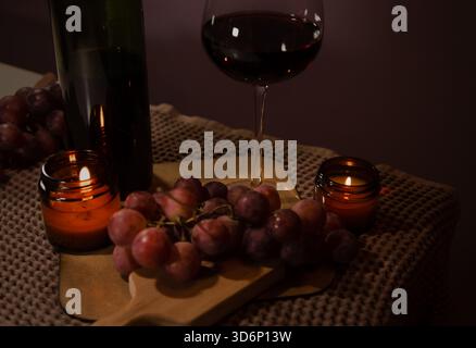Un bicchiere di vino da primo piano con vino rosso con bottiglia sul tavolo, grappoli di uva vicino a romantiche candele di fuoco fiammeggianti. Concetto di arredamento di Cristo Foto Stock