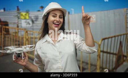 Ingegnere donna che tiene in mano un drone bianco e punta il dito indice verso la costruzione di barriere con elmetto visibile; eccitazione innovazione. Foto Stock