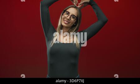 Donna con occhiali e lunghi capelli biondi forma il cuore con le mani, sorride e solleva le braccia contro un muro rosso profondo; gioia amore. Foto Stock