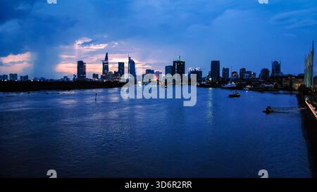 Vibrante paesaggio serale della città di ho chi Minh con grattacieli illuminati che si riflettono sul fiume Saigon sotto un suggestivo cielo blu al tramonto. Foto Stock
