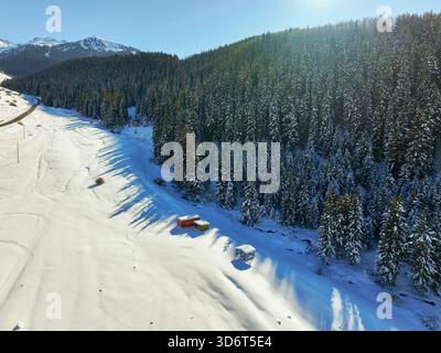 Veduta aerea di una fitta foresta di conifere che confina con un'ampia radura innevata con alcune piccole strutture colorate temporanee alla base dello slo Foto Stock