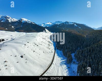 Vista panoramica aerea di una strada di montagna che si snoda tra un ampio campo innevato e una fitta foresta di conifere scura sotto un cielo azzurro Foto Stock