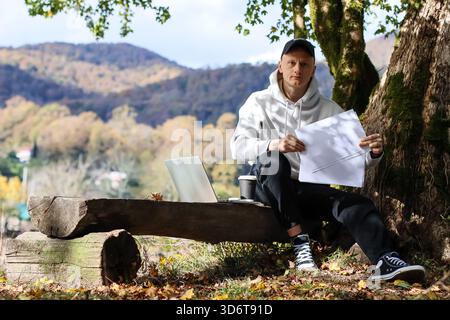 Mostrando un documento vuoto, un uomo siede su una panchina rustica in una foresta autunnale di montagna, il suo schermo del computer si illumina con un laptop aperto e una tazza di Foto Stock