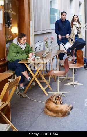 Parigi, Francia - marzo 2025: Una donna siede a un tavolino a mangiare, mentre una coppia con un passeggino si trova nelle vicinanze. Un cane si rilassa per terra all'esterno di Foto Stock