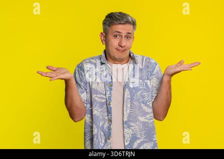 Uomo di mezza età che si scaglia le spalle e alza le mani con un aspetto incerto, esprimendo confusione e mancanza di risposte. Giovane ragazzo che mostra emozioni perplesse e reazioni poco chiare isolato su sfondo giallo Foto Stock