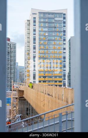 Cantiere con ponteggi in legno, un lavoratore cammina lungo le tavole. Sullo sfondo, alti edifici residenziali sorgono sopra il urb Foto Stock