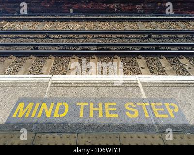 L'iconico cartello di sicurezza giallo "Mind the Step" dipinto sul bordo di un binario della stazione ferroviaria, che consiglia ai passeggeri di prestare attenzione vicino al binario. Foto Stock