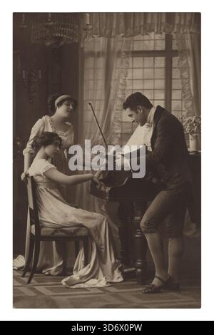 Una cartolina vintage dai toni seppia mostra tre persone in un salotto; un uomo suona il violino, una donna siede ad ascoltare un pianoforte e un'altra donna sta Foto Stock