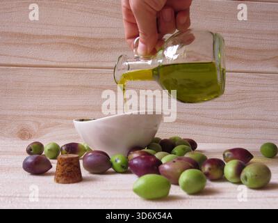Olio d'oliva naturale ed ecologico in Grecia versato in un piccolo piatto a mano, circondato da olive verdi e nere fresche sul tavolo. Foto Stock