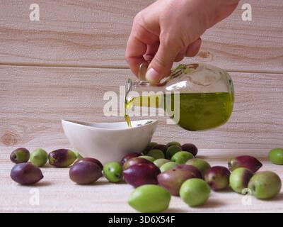 Olio d'oliva naturale ed ecologico in Grecia versato in un piccolo piatto a mano, circondato da olive verdi e nere fresche sul tavolo. Foto Stock