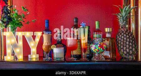 Cocktail colorati, bottiglie di liquori, caramelle ed elementi decorativi disposti su un tavolino da bar festivo con vibrante illuminazione di sfondo rosso. Foto Stock
