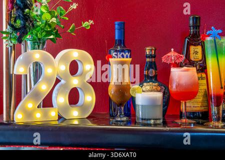 Cocktail colorati, bottiglie di liquori, caramelle ed elementi decorativi disposti su un tavolino da bar festivo con vibrante illuminazione di sfondo rosso. Foto Stock