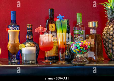 Cocktail colorati, bottiglie di liquori, caramelle ed elementi decorativi disposti su un tavolino da bar festivo con vibrante illuminazione di sfondo rosso. Foto Stock
