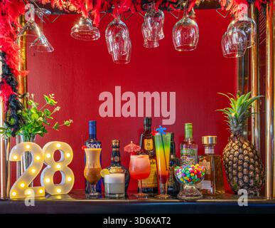 Cocktail colorati, bottiglie di liquori, caramelle ed elementi decorativi disposti su un tavolino da bar festivo con vibrante illuminazione di sfondo rosso. Foto Stock
