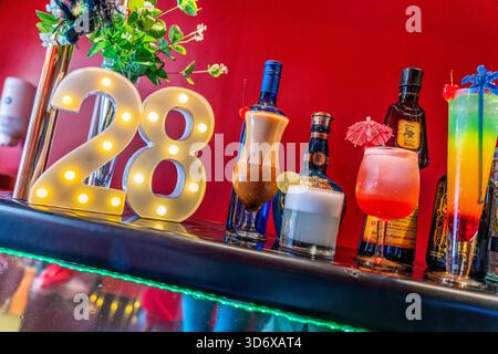 Cocktail colorati, bottiglie di liquori, caramelle ed elementi decorativi disposti su un tavolino da bar festivo con vibrante illuminazione di sfondo rosso. Foto Stock
