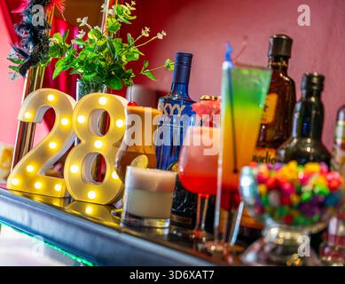 Cocktail colorati, bottiglie di liquori, caramelle ed elementi decorativi disposti su un tavolino da bar festivo con vibrante illuminazione di sfondo rosso. Foto Stock