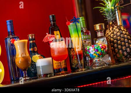 Cocktail colorati, bottiglie di liquori, caramelle ed elementi decorativi disposti su un tavolino da bar festivo con vibrante illuminazione di sfondo rosso. Foto Stock
