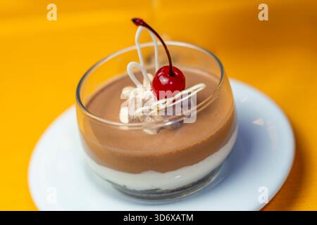 Dessert di panna cotta al cioccolato ricoperto di ciliegia, panna montata e decorazione di cioccolato bianco servito in una tazza di vetro su un piatto bianco su un coagulo giallo Foto Stock