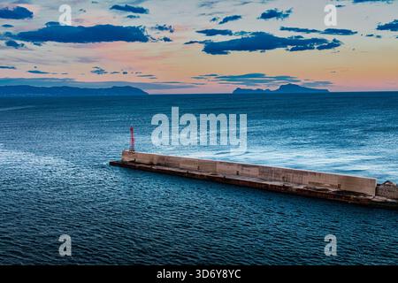 L'immagine mostra un lungo molo di cemento grigio che si estende nell'oceano. Il molo è affiancato da un'alta torre di comunicazione rossa. Una costa lontana, featu Foto Stock
