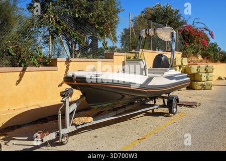 Un rimorchio con motoscafo gonfiabile è parcheggiato sulla strada contro un muro, con piante fiorite e blocchi modulari di pontoni sullo sfondo Foto Stock