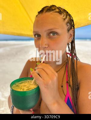 Una giovane donna in bikini con i capelli intrecciati si gode un cocktail in un vivace bicchiere verde su una spiaggia soleggiata. Foto Stock