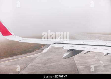Ala dell'aereo vista dal finestrino del passeggero mentre l'aeromobile opera sulla pista in fitta nebbia. La pista in calcestruzzo e la visibilità limitata creano un senso di umorismo Foto Stock