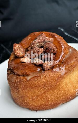 Primo piano di un rotolo di cannella glassato con noci candite su un piatto bianco su sfondo scuro Foto Stock