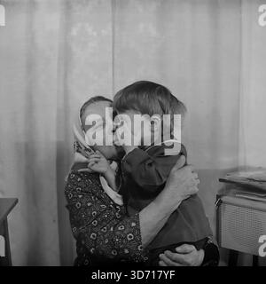 Slaviansk, regione di Donetsk, RSS Ucraina, Unione Sovietica. Febbraio 1986. Una toccante scena domestica che coinvolge la famiglia del vice Marchenko. L'anziana madre del vice, che indossa un tradizionale velo, tiene un bambino piccolo tra le braccia. La bambina si tocca scherzosamente con il viso e la abbraccia mentre sorride calorosamente. Sono a casa vicino a una radio d'epoca. Questa fotografia d'archivio in bianco e nero cattura il profondo legame tra generazioni nella pacifica regione del Donbass molto prima della guerra. Foto Stock