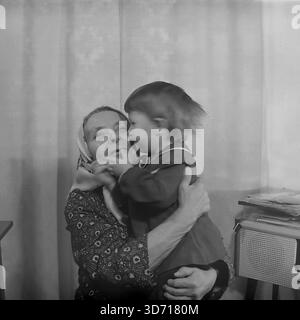 Slaviansk, regione di Donetsk, RSS Ucraina, Unione Sovietica. Febbraio 1986. Una toccante scena domestica che coinvolge la famiglia del vice Marchenko. L'anziana madre del vice, che indossa un tradizionale velo, tiene un bambino piccolo tra le braccia. La bambina si tocca scherzosamente con il viso e la abbraccia mentre sorride calorosamente. Sono a casa vicino a una radio d'epoca. Questa fotografia d'archivio in bianco e nero cattura il profondo legame tra generazioni nella pacifica regione del Donbass molto prima della guerra. Foto Stock