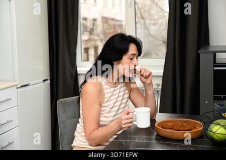 Una donna russa matura di 50 anni si gode una mattinata tranquilla e sonnolenta tenendo un caffè e un biscotto, prendendo un boccone mentre si siede tranquillamente con un morbido e pacifico m Foto Stock