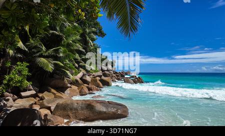 Acqua turchese cristallina che si bagna su una spiaggia sabbiosa con lisce rocce di granito e un'isola lontana all'orizzonte. Seychelles, Praslin. Anse Georgette. Foto Stock