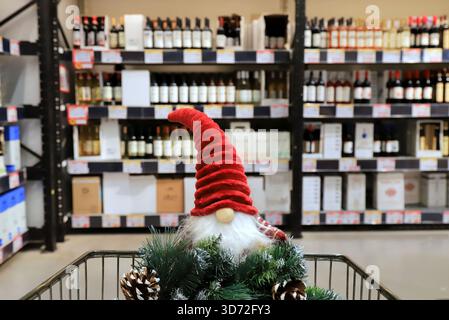 Divertente gnomo natalizio e corona su un carrello da supermercato. Carrello della spesa, scaffali con bottiglie di alcol, cibo, alimentari. Shopping in negozio, Capodanno Foto Stock