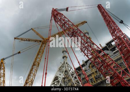 Una massiccia gru cingolata con bracci a reticolo rossi posizionata in un cantiere di raffineria, che solleva attrezzature vicino a torri di processo alte sotto lo sci nuvoloso Foto Stock