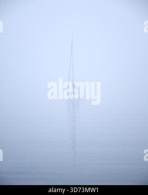Una barca a vela solitaria galleggia tranquillamente sull'acqua calma, circondata da una fitta nebbia. La scena sembra tranquilla, misteriosa e tranquilla, con contorni morbidi e generosità Foto Stock