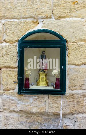 Piccolo Santuario religioso esposto dietro il vetro nella finestra ad arco incastonata nella calda parete di pietra Foto Stock