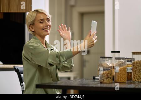 Donna caucasica di mezza età sorridente e saluta la mano tenendo lo smartphone, seduto al tavolo della cucina con vari contenitori di cibo, impegnato in videochiamate o conversazione online Foto Stock