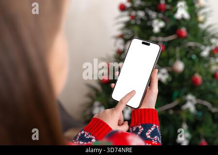 Ragazza che tiene in mano uno smartphone con schermo vuoto vicino all'albero di Natale, perfetta per i mockup delle app per le vacanze, la pubblicità stagionale, le promozioni mobili e le festività Foto Stock