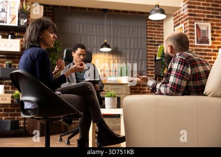 La donna ansiosa sembra sconvolta parlando con il marito irritato durante la sessione di terapia mentre lo psichiatra maschile nero scrive delle note. Coppia sposata che lotta per la consulenza, discute di lotte emotive. Foto Stock
