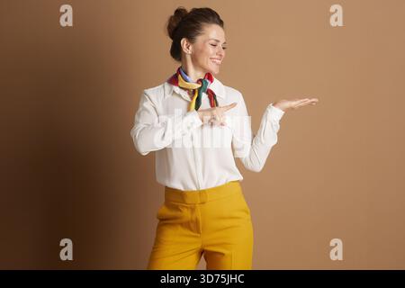 Donna professionista sorridente con camicia bianca, sciarpa colorata e pantaloni gialli, punti e gesti, che presenta informazioni o mette in evidenza un'opzione Foto Stock