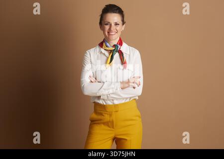 Una donna d'affari sorridente con una sciarpa colorata e pantaloni gialli sta con le braccia incrociate, trasudando fiducia, stile e prontezza professionale Foto Stock
