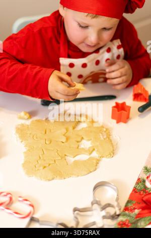 Un bambino con un vestito rosso taglia con gioia le forme dei biscotti dall'impasto a un tavolo decorato per le vacanze. I colori brillanti e gli strumenti per le feste creano un'allegria Foto Stock