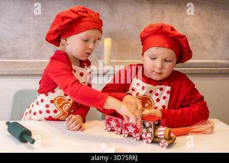 Due bambini in cappelli rossi da chef e grembiuli cucinano insieme in una cucina moderna. Decorano dolcetti e condividono momenti di gioia. L'atmosfera Foto Stock