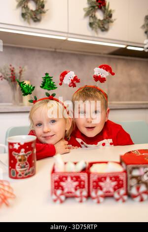 Due bambini con giocosi fasciatesta per le vacanze posano insieme in una cucina luminosa. Sono circondate da decorazioni festose mentre si godono il Natale Foto Stock