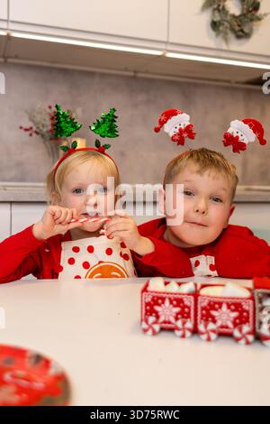 Due bambini che indossano cerchietti festivi stanno preparando prelibatezze natalizie in una cucina luminosa. Si godono il momento, mostrando gioia e creatività durante i festeggiamenti Foto Stock