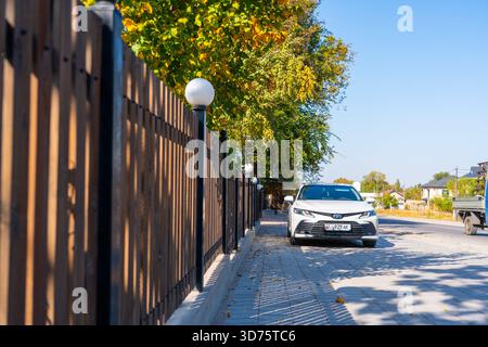 Talas, Kazakistan - 13 settembre 2024: Una berlina Toyota Camry bianca parcheggiata su un marciapiede soleggiato accanto a una recinzione di legno e luci a globo, con un colle autunnale Foto Stock