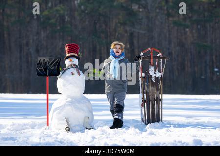 I bambini giocano con pupazzo di neve e slitta. Giornata invernale attiva per i bambini. Infanzia in inverno. Slitta per bambini e pupazzo di neve. Vacanze invernali. Bambino con gioco di slitta Foto Stock