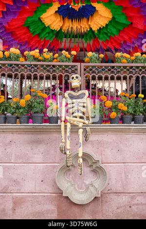 Uno scheletro è seduto su una sporgenza di fronte a un arco colorato. L'arco è decorato con fiori. Messico, zacatlan de las manzanas, puebla, giorno Foto Stock