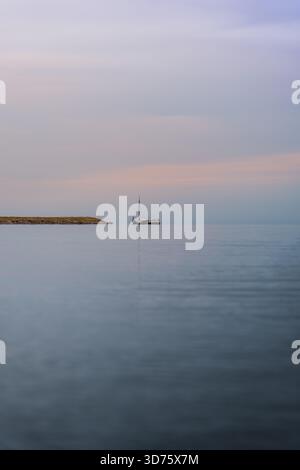 La barca a vela Dutch Tjalk si allontana pacificamente sull'acqua piatta vicino alla riva, con un cielo pastello leggermente nuvoloso al crepuscolo. La scena cattura la serenità serale. Vista ad angolo basso Foto Stock