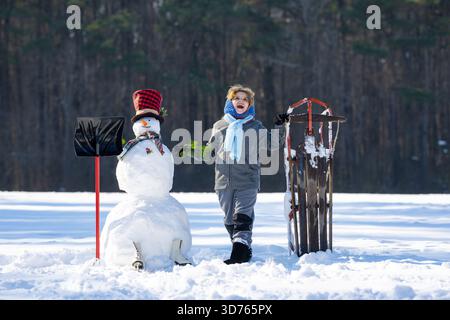 I bambini giocano con pupazzo di neve e slitta. Giornata invernale attiva per i bambini. Infanzia in inverno. Slitta per bambini e pupazzo di neve. Vacanze invernali. Bambino con gioco di slitta Foto Stock